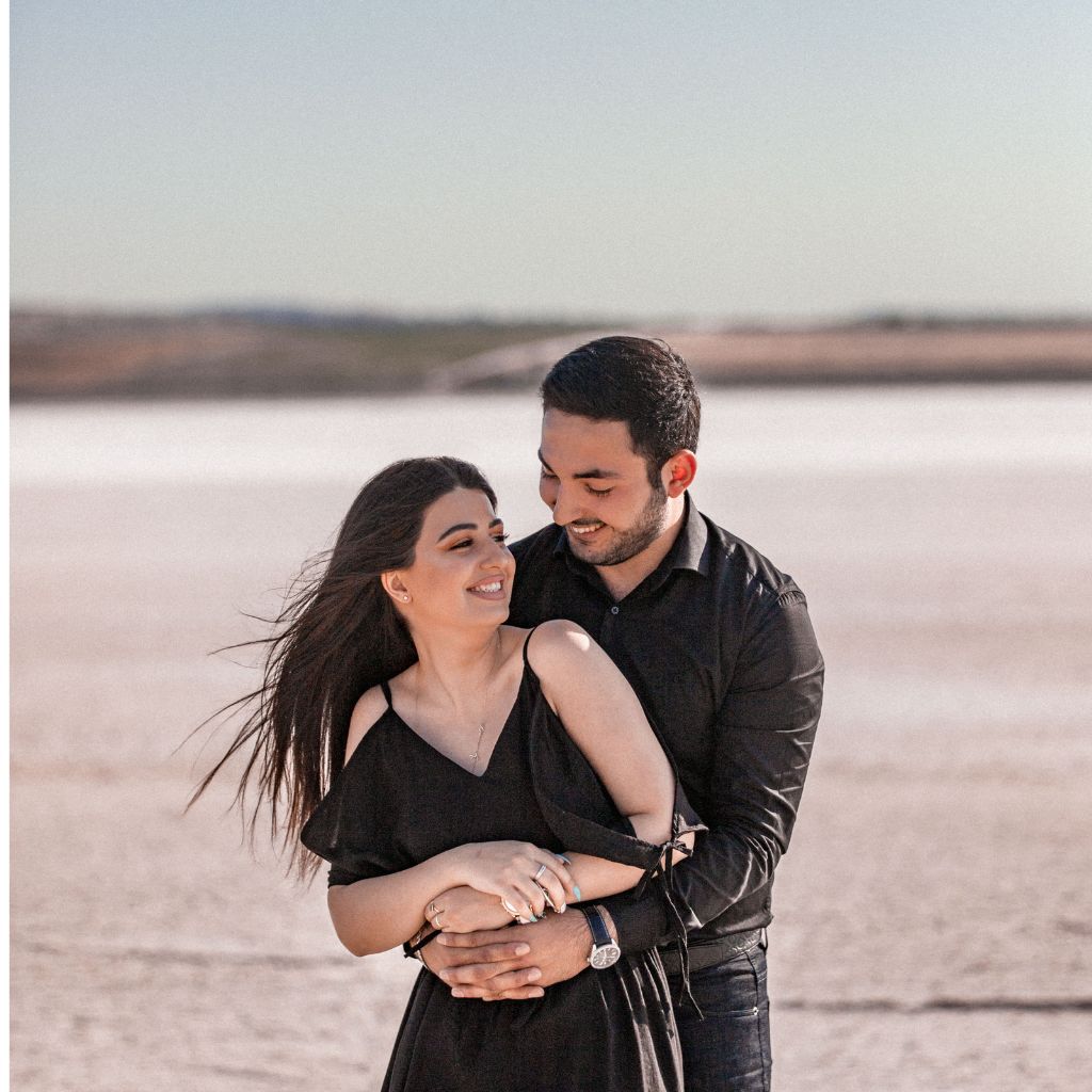 Smiling couple embracing outdoors near a calm landscape, with the man standing behind the woman.